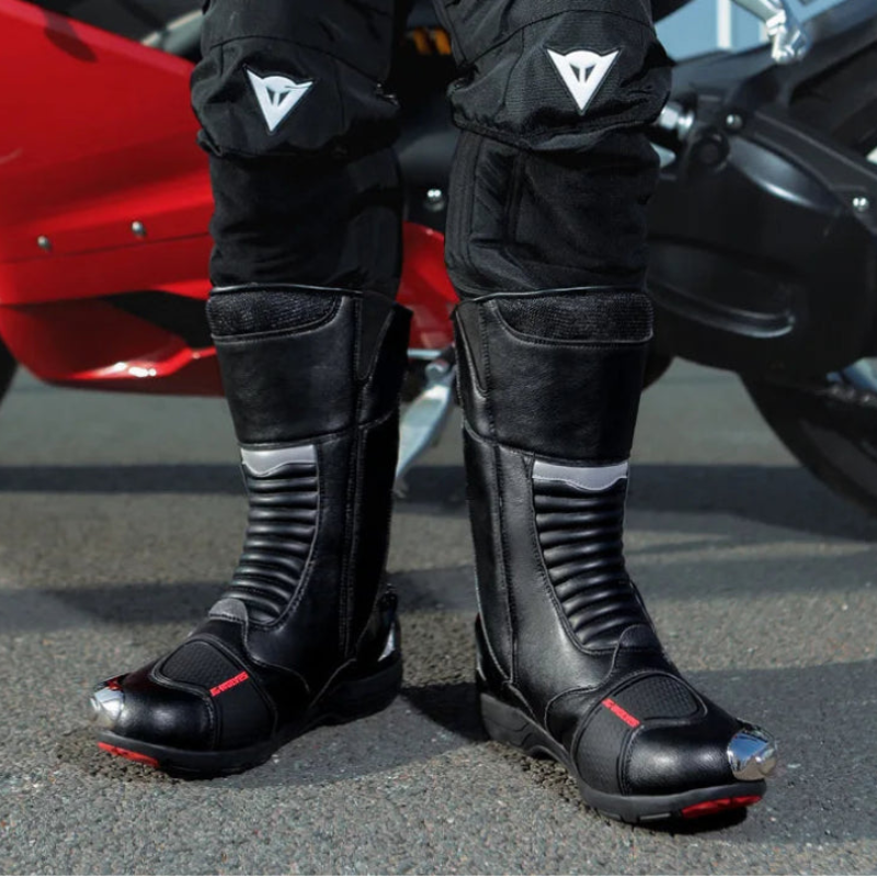 Person wearing black motorcycle boots and pants with visible brand logo, standing next to a red motorcycle.