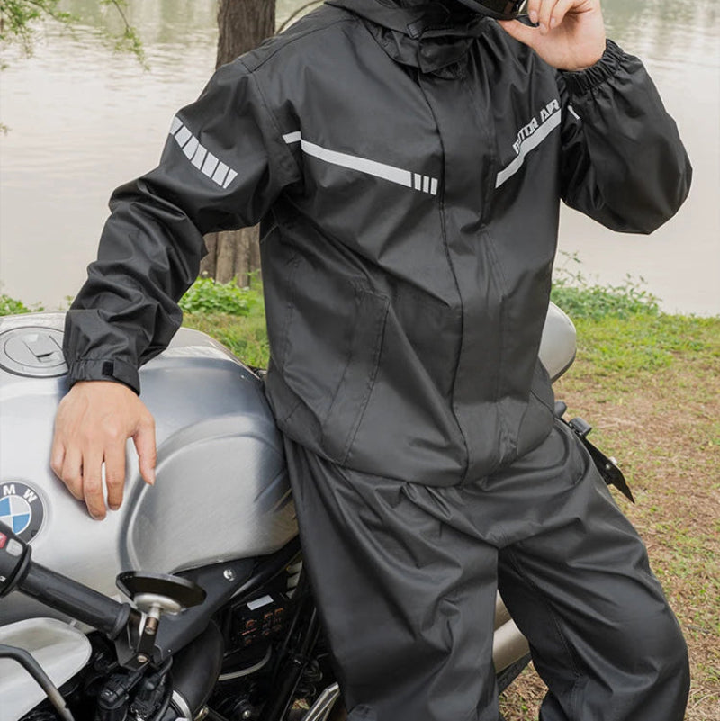 Person wearing a black rain suit and helmet next to a motorcycle by a lake.